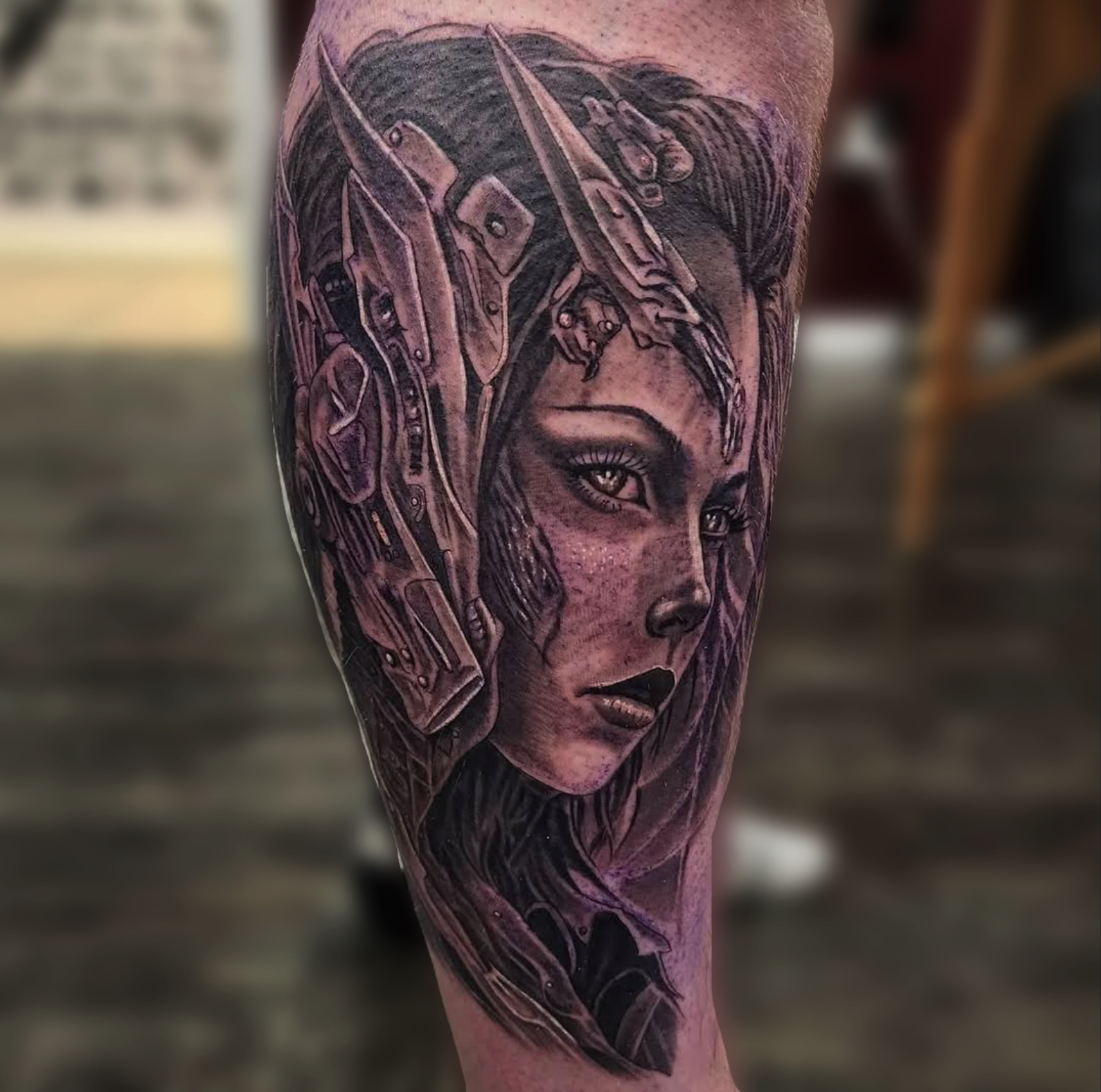 Black and grey realism calf tattoo of a woman's face framed by biomechanical armour and blades