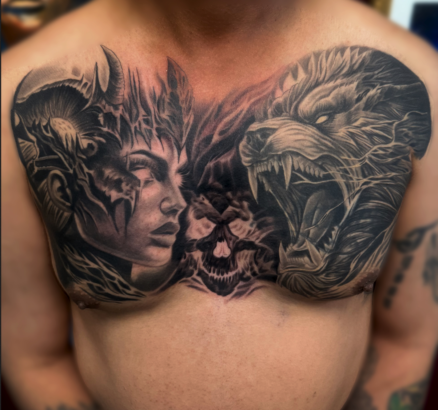 Black and grey realism chest piece of a horned woman facing a snarling wolf with a skull between them